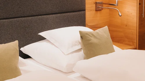 Service im Hotel Panorama in Lech am Arlberg Hotelbett mit weißer Bettwäsche, beigem Zierkissen, gepolstertem Kopfteil und Wandlampe