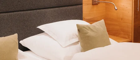 Hotel bed with white linens, a beige accent pillow, padded headboard, and wall lamp