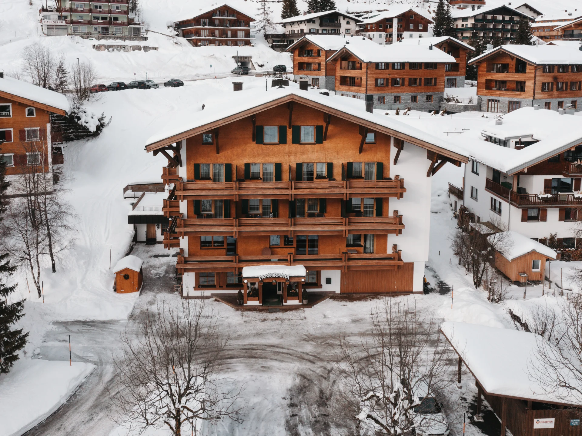 The journey to your hotel in Vorarlberg | Hotel Panorama Wooden chalet with balconies in a snowy mountain village, surrounded by other buildings
