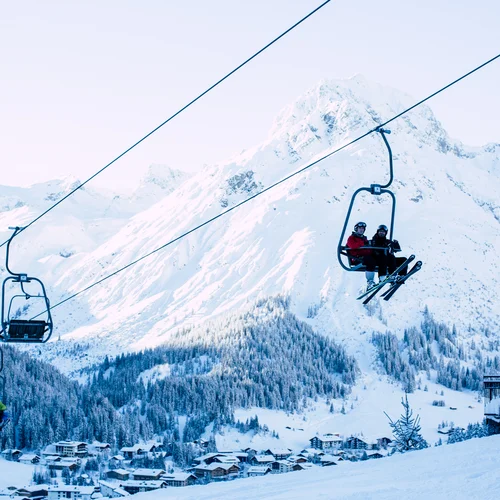 Inclusive services | Hotel Panorama in Lech am Arlberg Ski lift carrying skiers past a snowy mountain and a village in the valley