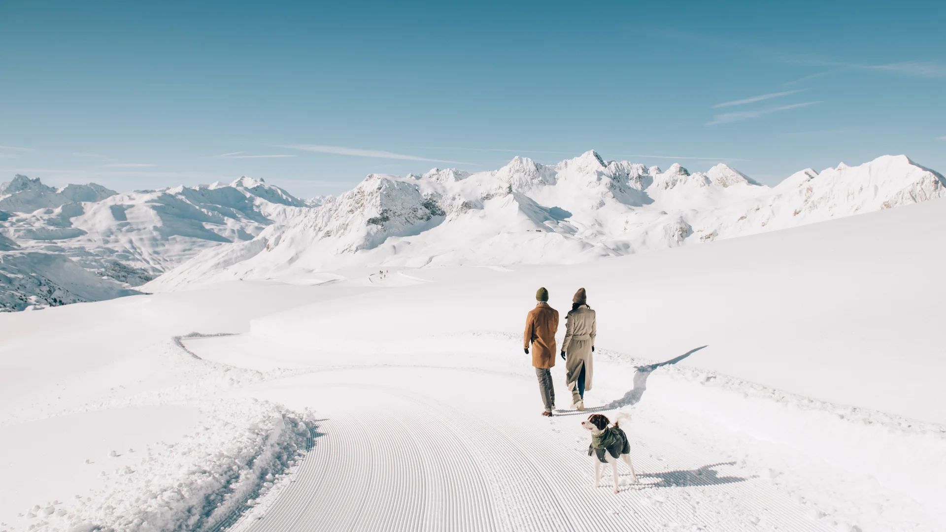 Zwei Personen mit Hund spazieren auf einem präparierten Schneepfad vor verschneiten Berggipfeln