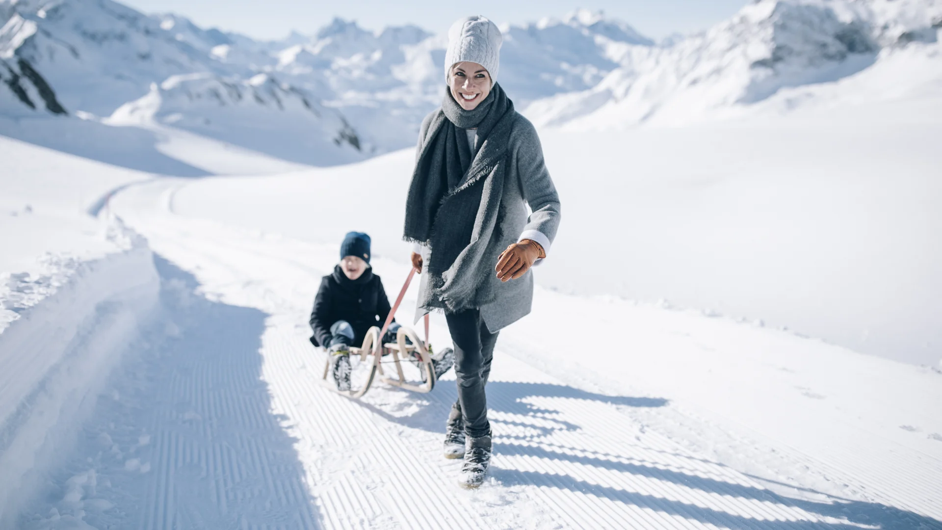 Frau und Kind mit Schlitten auf einem präparierten Winterweg vor verschneiten Bergen