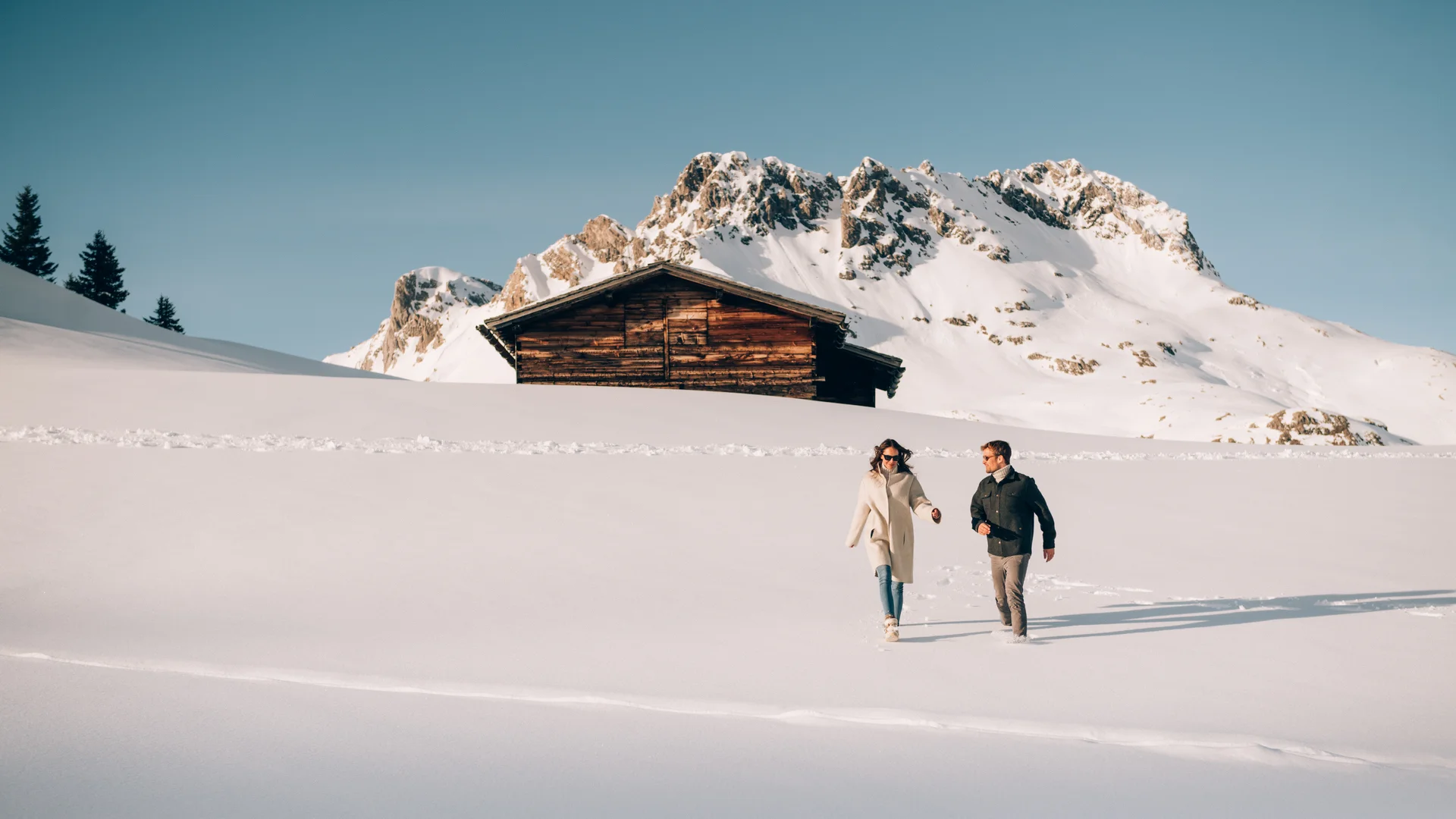 Picture gallery | Hotel Panorama in Lech am Arlberg Two people walk through a snowy landscape with a mountain hut and snow-covered peaks