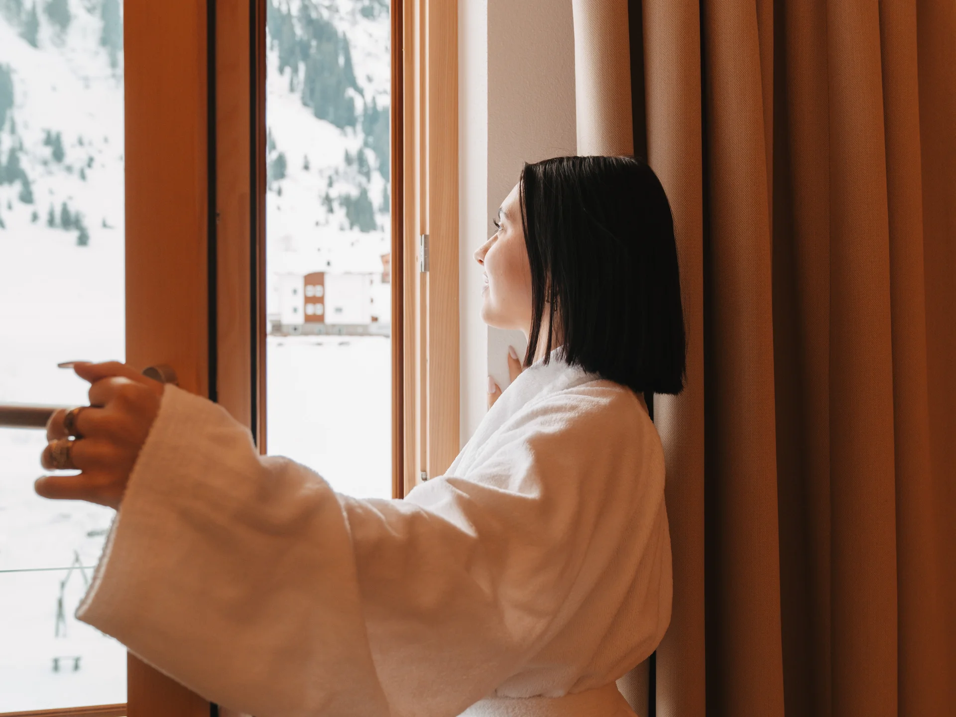 Frau im weißen Bademantel blickt aus dem Fenster auf eine verschneite Berglandschaft