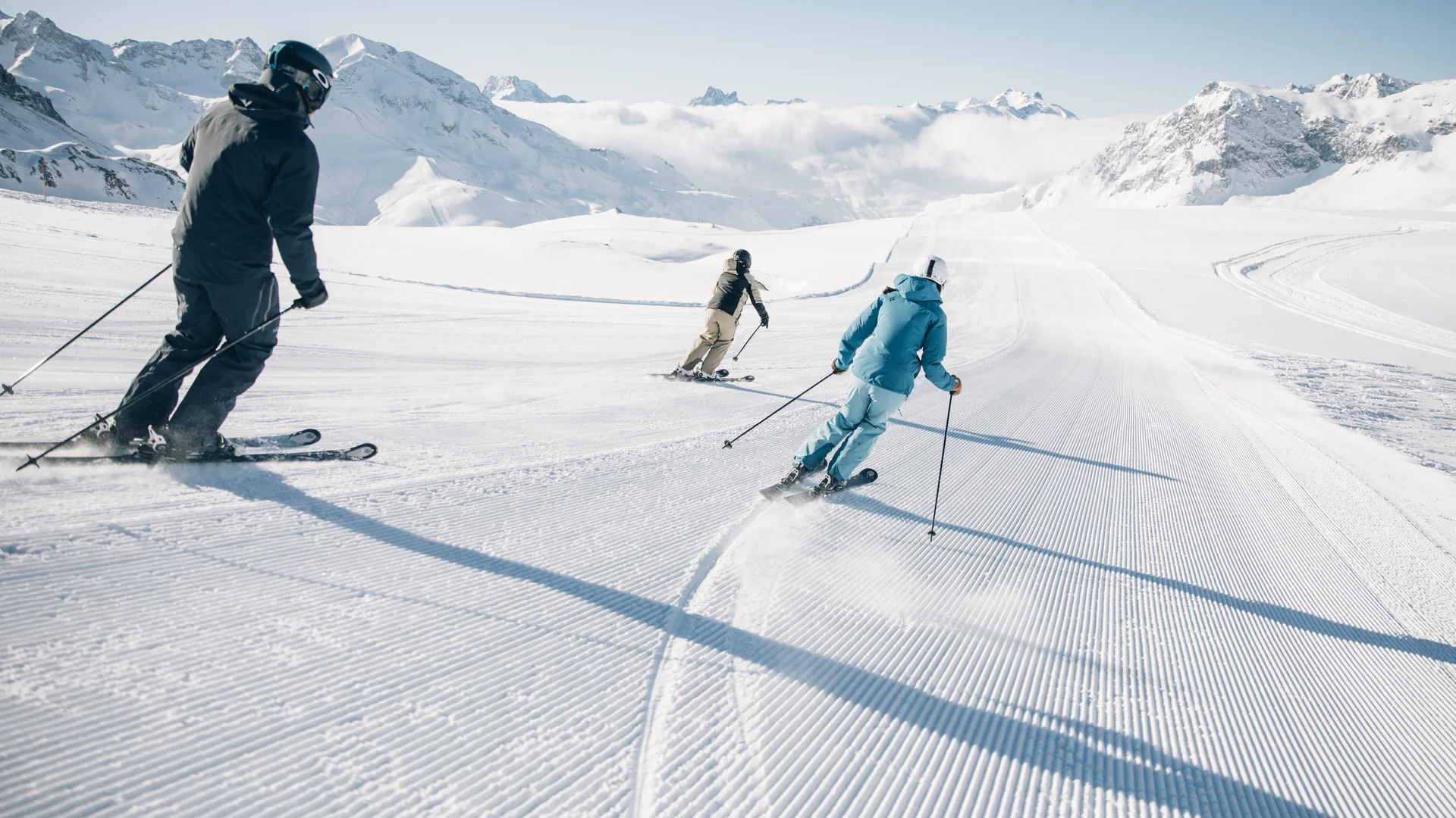 Picture gallery | Hotel Panorama in Lech am Arlberg Three skiers descend a groomed slope with snow-covered mountains in the background