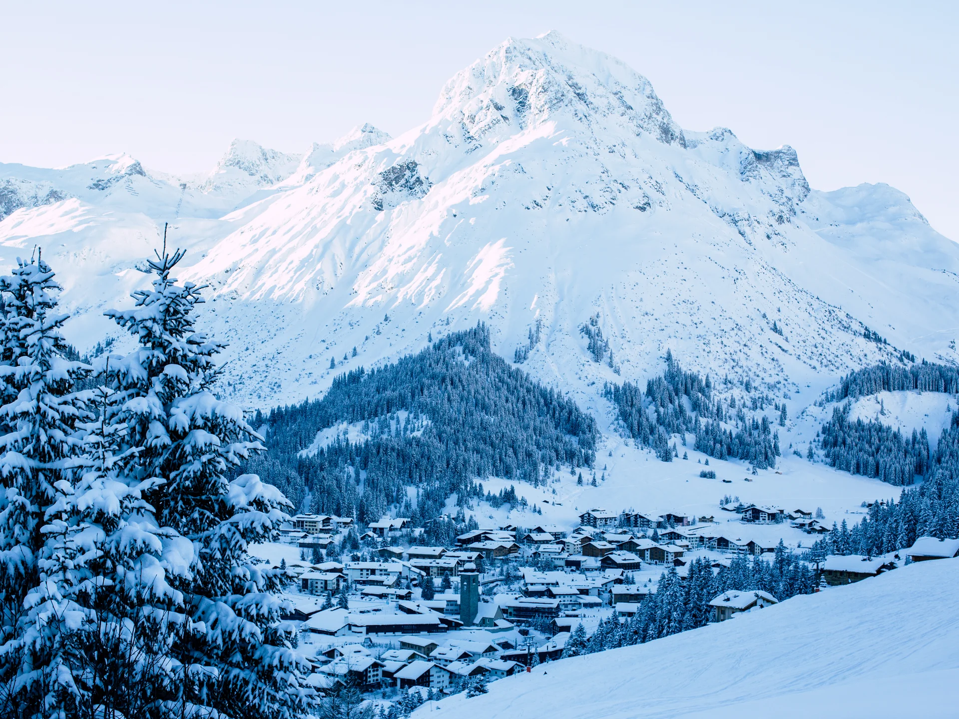The journey to your hotel in Vorarlberg | Hotel Panorama Snow-covered mountain village beneath a towering peak, surrounded by forests and snowy fir trees